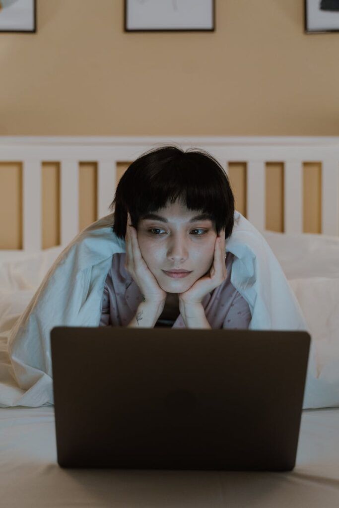 A woman comfortably browsing on her laptop while lying in bed, under a warm blanket.