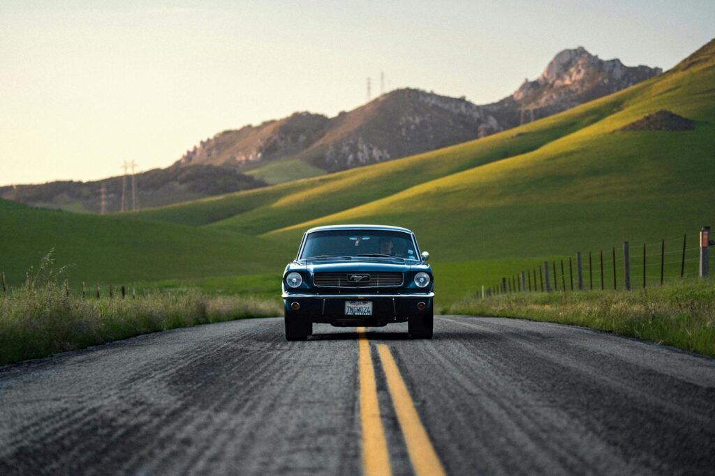 Old-fashioned car driving on a rural road with scenic green hills in the background.