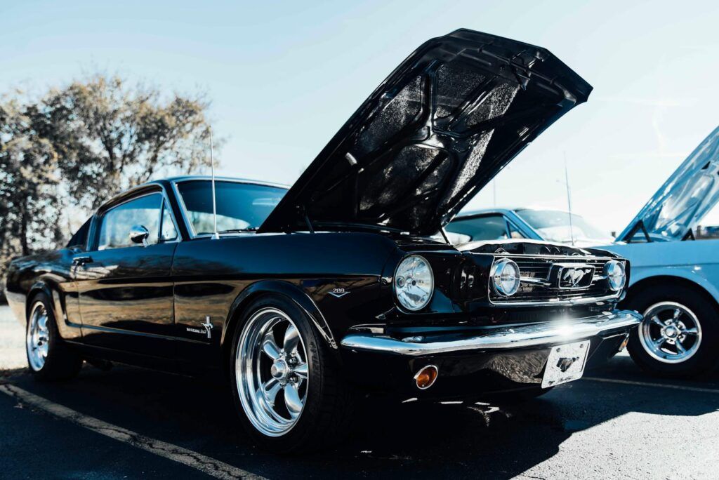 Vintage black Mustang with hood up parked outdoors on a sunny day.