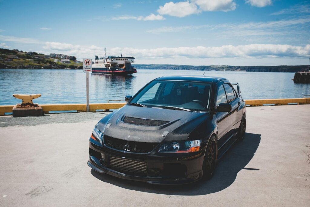 Black Mitsubishi Lancer Evolution parked by a scenic harbor with a ferry in the background.