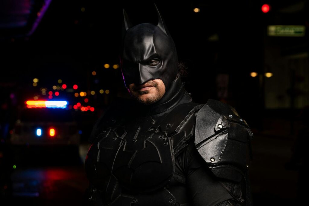 Batman cosplayer in detailed costume at a night event with police lights in the background.