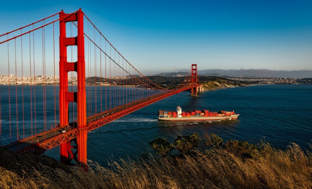 Golden Gate Bridge arches over San Francisco Bay as a cargo ship passes beneath during sunset.