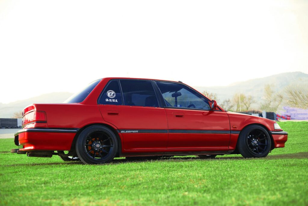 A striking red Honda Civic sedan with custom modifications parked on a green field.