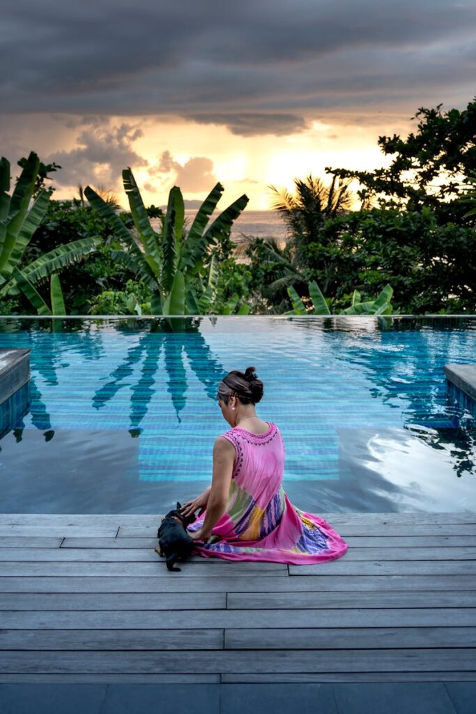 A woman and her black dog enjoy a serene sunset by a tropical infinity pool.