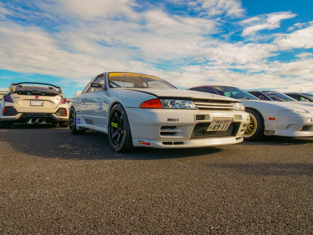 A vibrant outdoor car meet showcasing iconic JDM cars including a Nissan Skyline and Honda Civic.