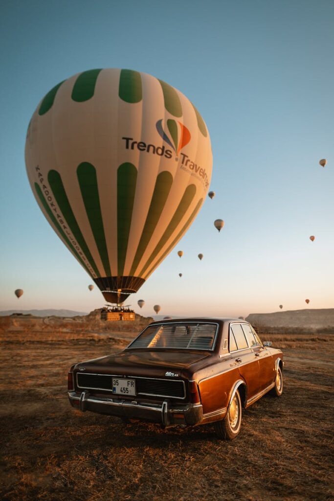 Classic car parked with hot air balloons in Cappadocia, Türkiye at sunrise.