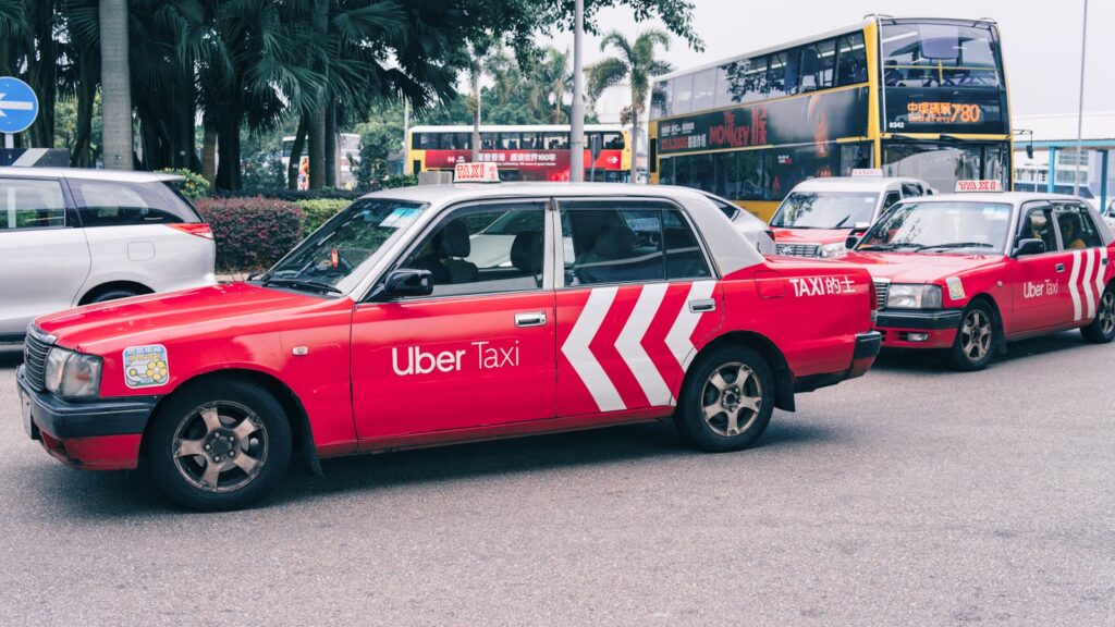 Red Uber taxi parked on a bustling city street with buses and greenery in the background.