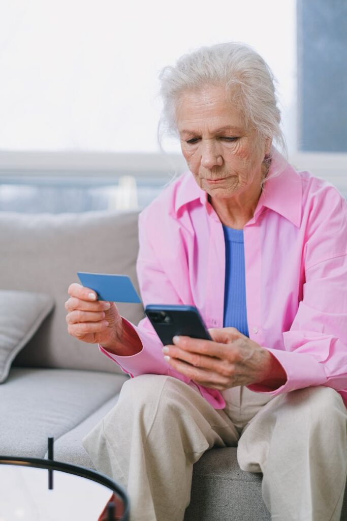 Senior woman using a credit card and smartphone for online shopping in a bright living room.