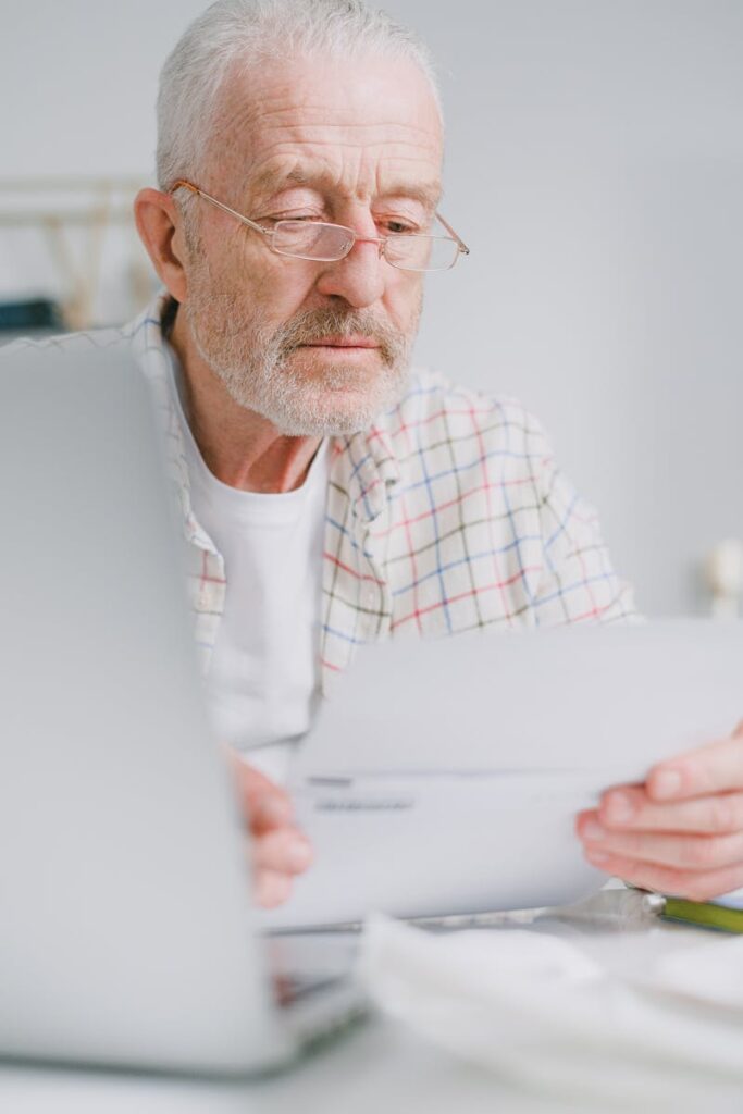 Elderly man with glasses reviews paperwork while sitting indoors with a laptop.