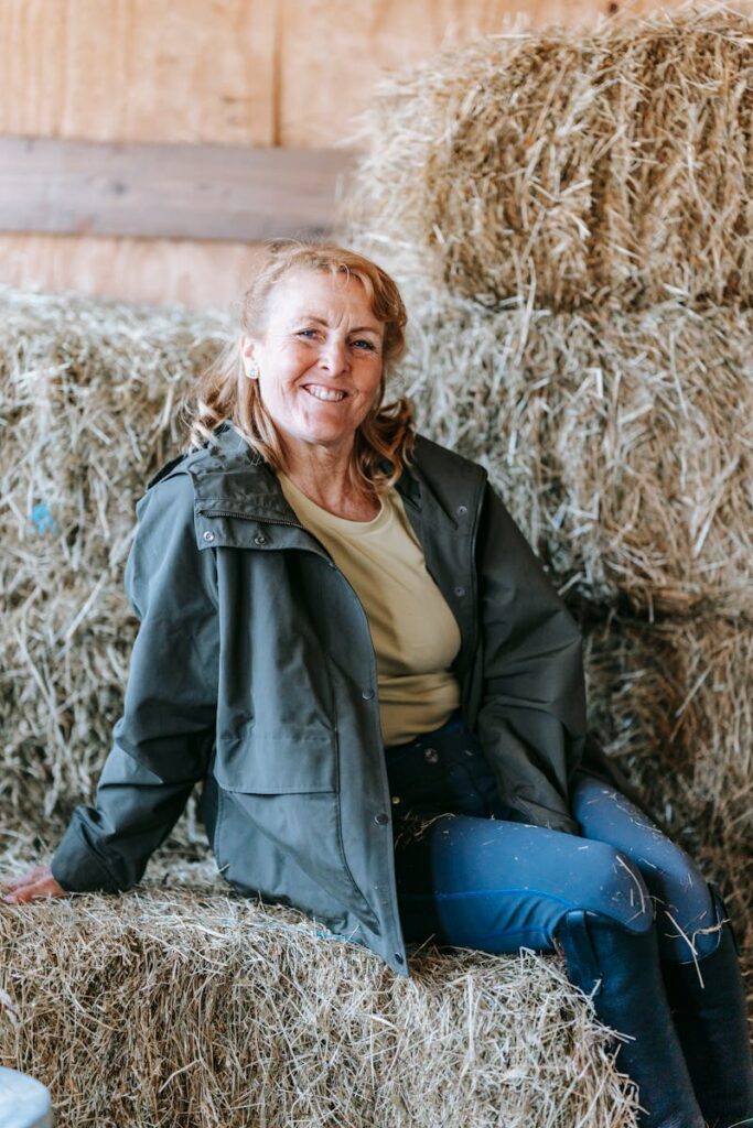 Smiling woman in an outdoor jacket seated on hay bales, exuding warmth and rustic charm.