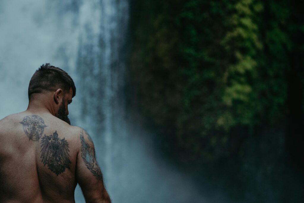 A shirtless man with tattoos stands by a waterfall, showcasing strength in Cascade Locks, OR.