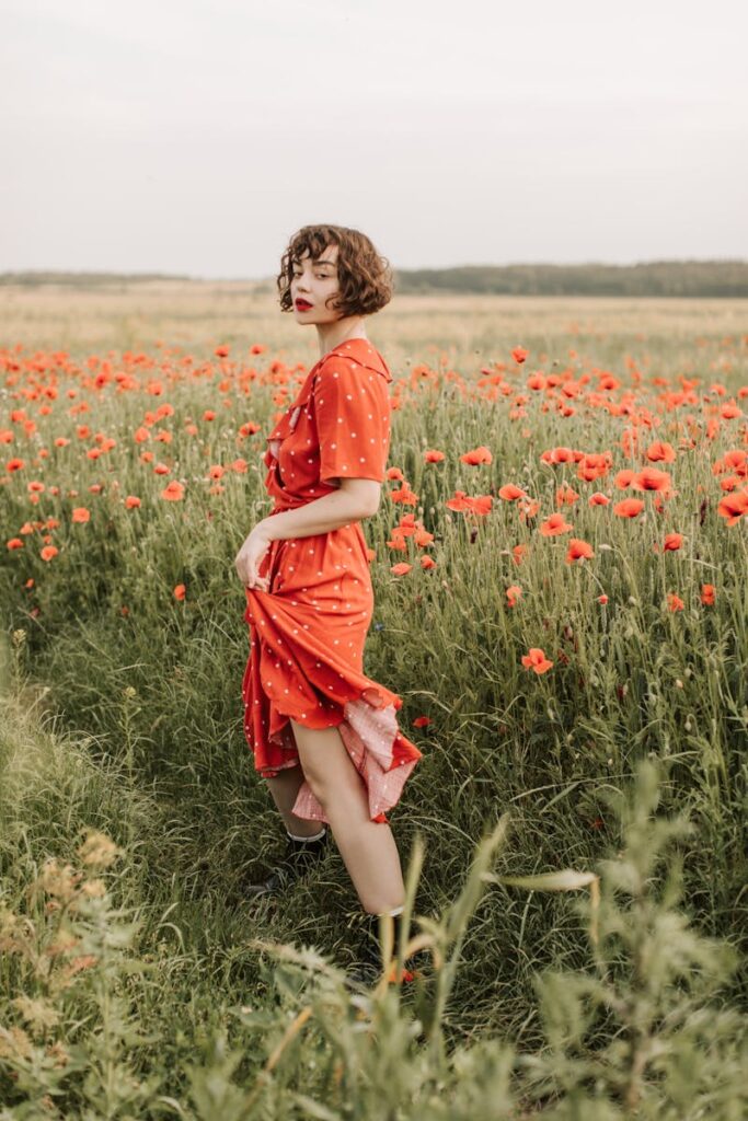 A woman in a red dress poses gracefully in a vibrant poppy field during the day.