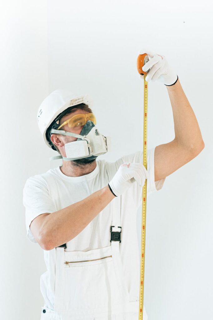 A construction worker in full safety gear measures with a tape against a white wall.