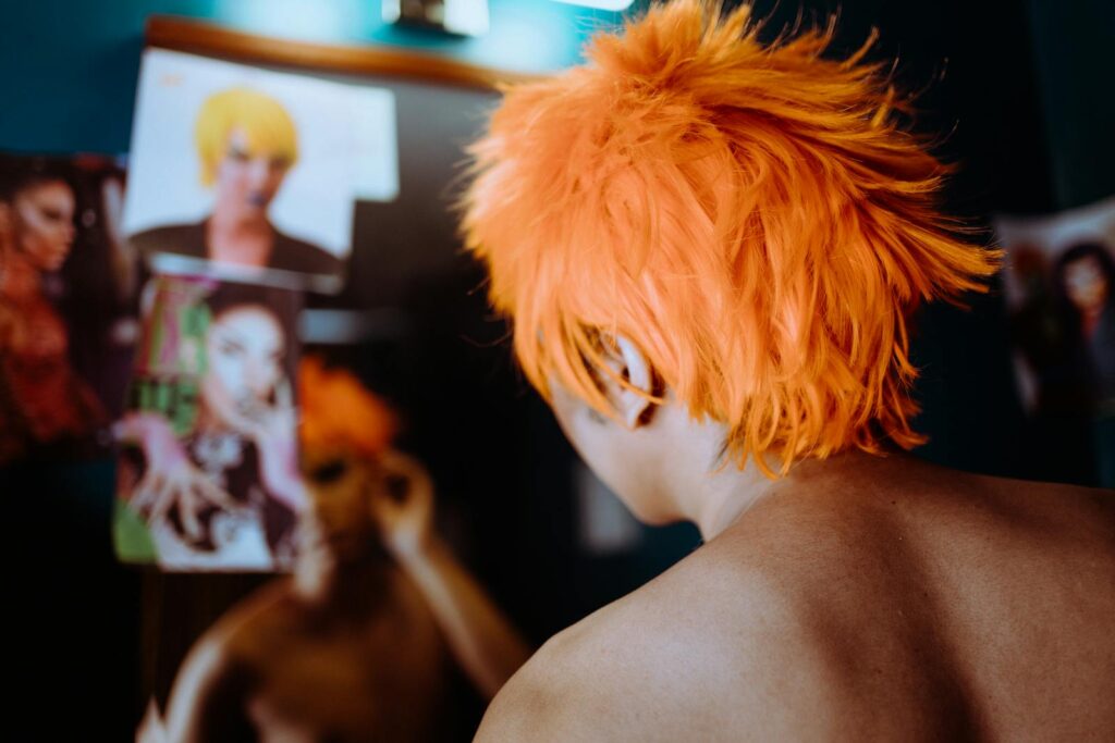 Back view of anonymous ethnic naked androgynous male stylist looking at mirror under LED lamp