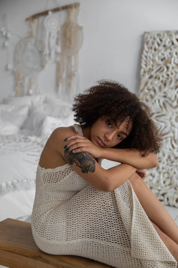 Side view of sad African American female embracing knees while sitting in cozy bedroom