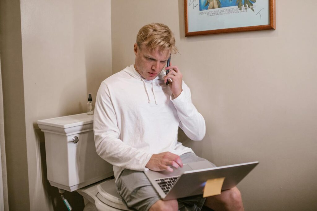 A man multitasks by talking on the phone and using a laptop while sitting in a bathroom.