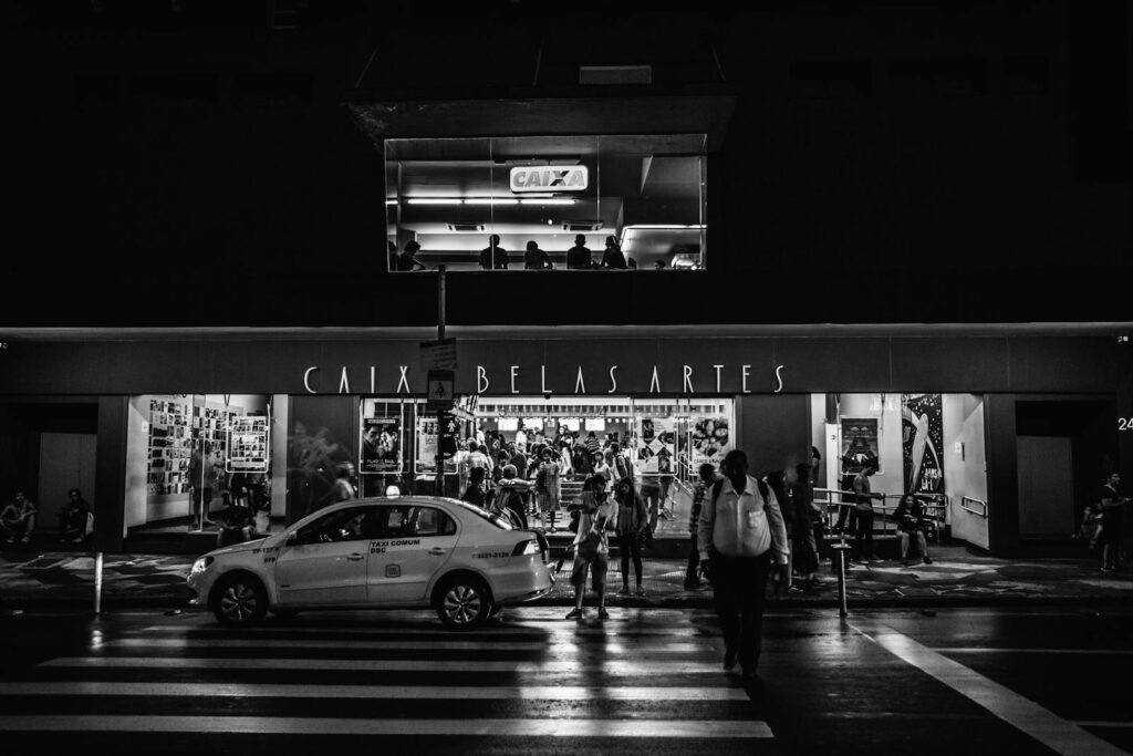 Black and white image of Caixa Belas Artes cinema entrance with people and a taxi at night.
