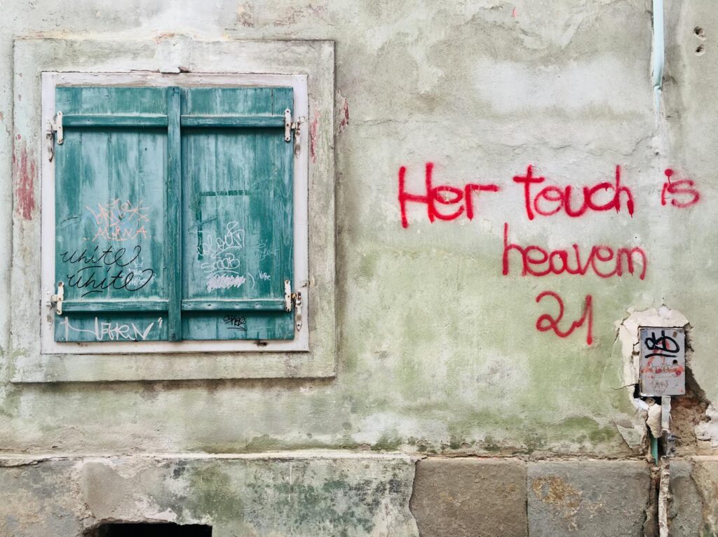 Old green shuttered window with graffiti and red text on wall in Brașov, Romania.