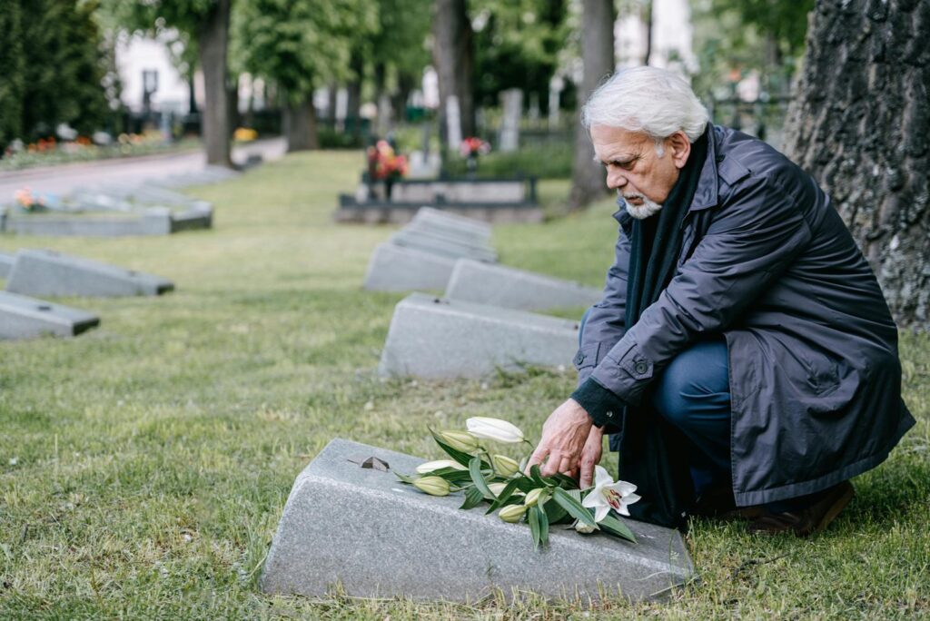 Senior man with white hair honorably placing lilies on a grave in a peaceful cemetery setting.