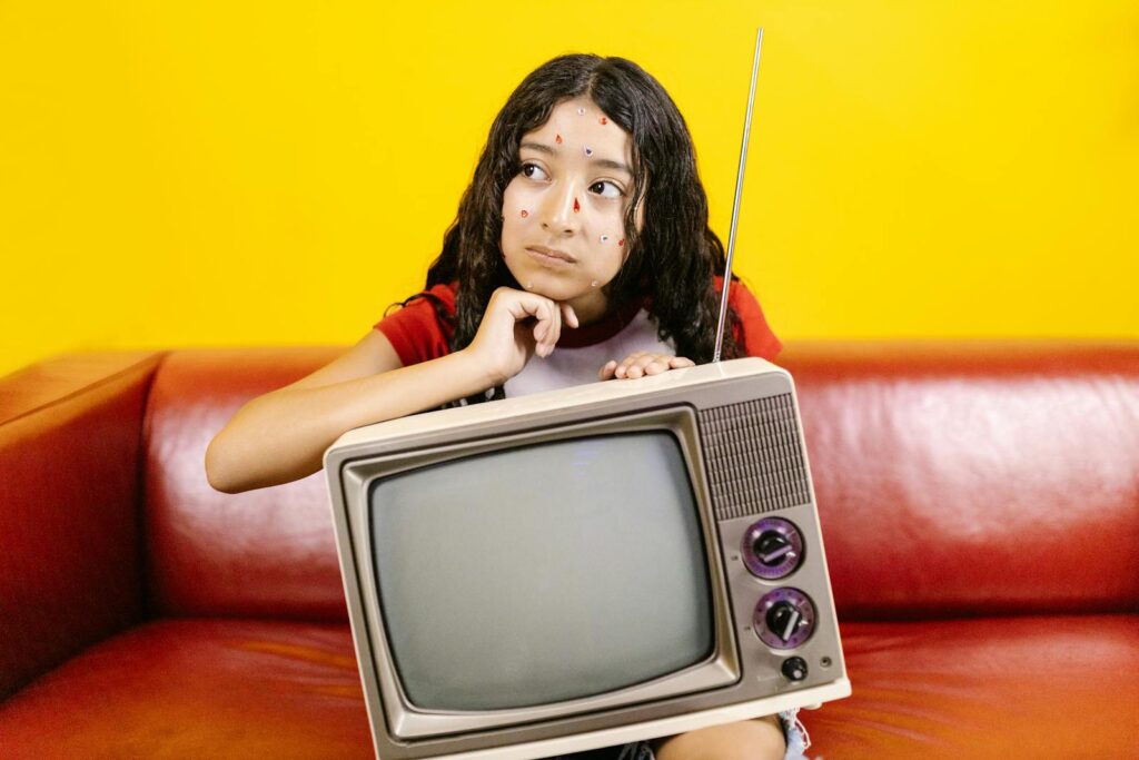 Woman with stickers on face posing with a vintage TV on a red couch with a bright yellow background.