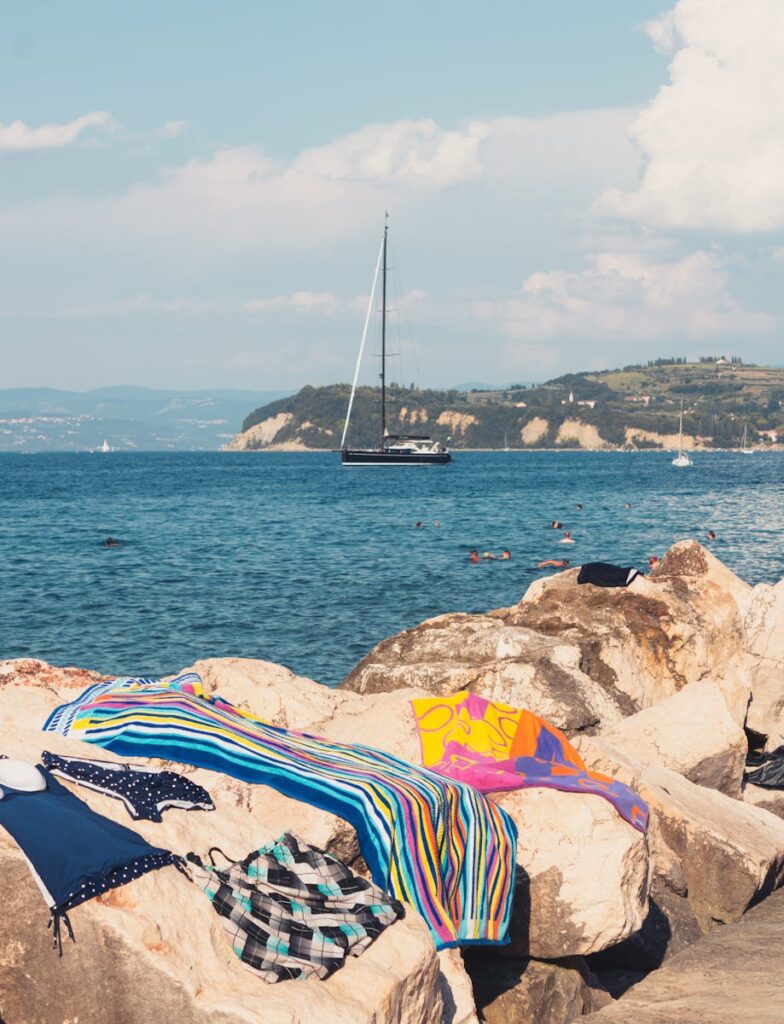 Beach towels on rocks with a sailboat in the distance on a bright day in Piran, Slovenia.