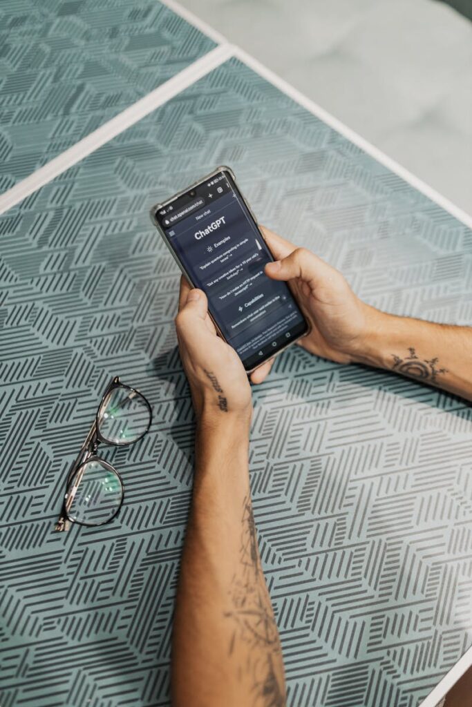 Hands holding smartphone displaying ChatGPT app near eyeglasses on table.