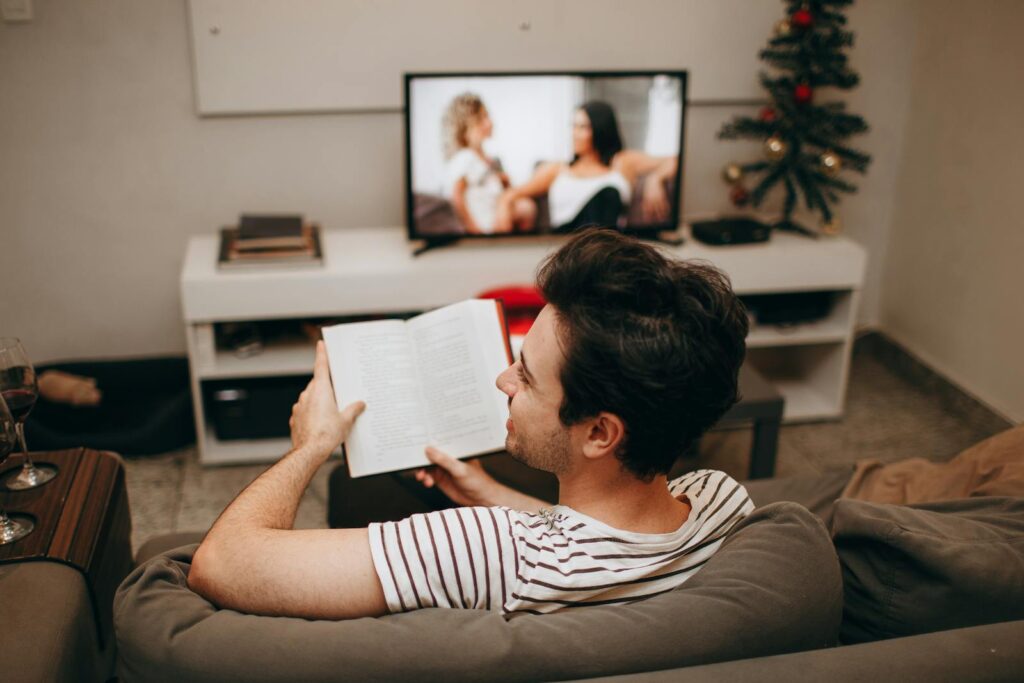 A man sits on a sofa reading a book in a cozy living room with a TV on.