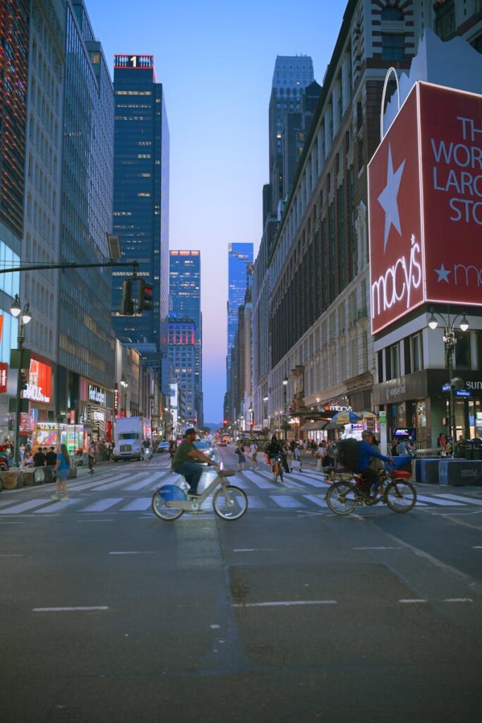 Bicyclists crossing a bustling intersection near Macy's in New York City during twilight.