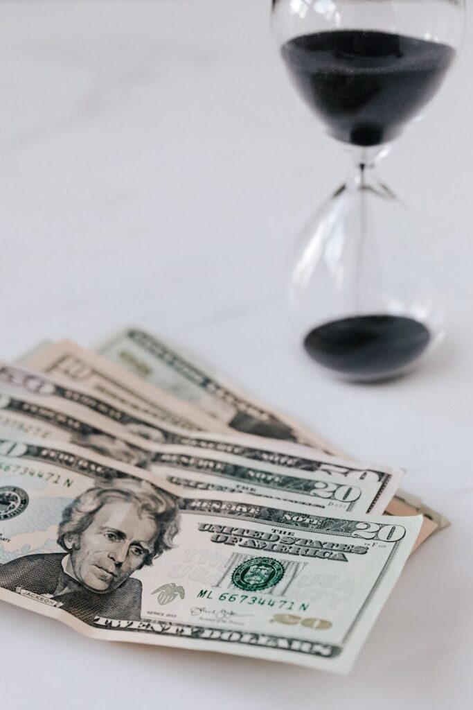 From above of transparent hourglass with black sand and heap of American dollars with image of president
