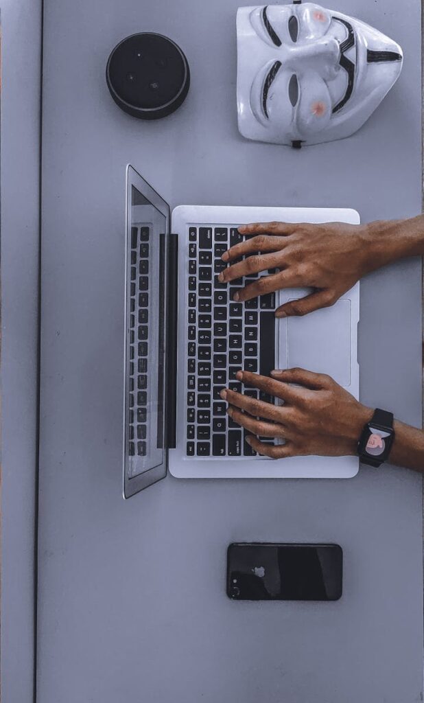 Top view of hands typing on a laptop beside a mask and smartphone.