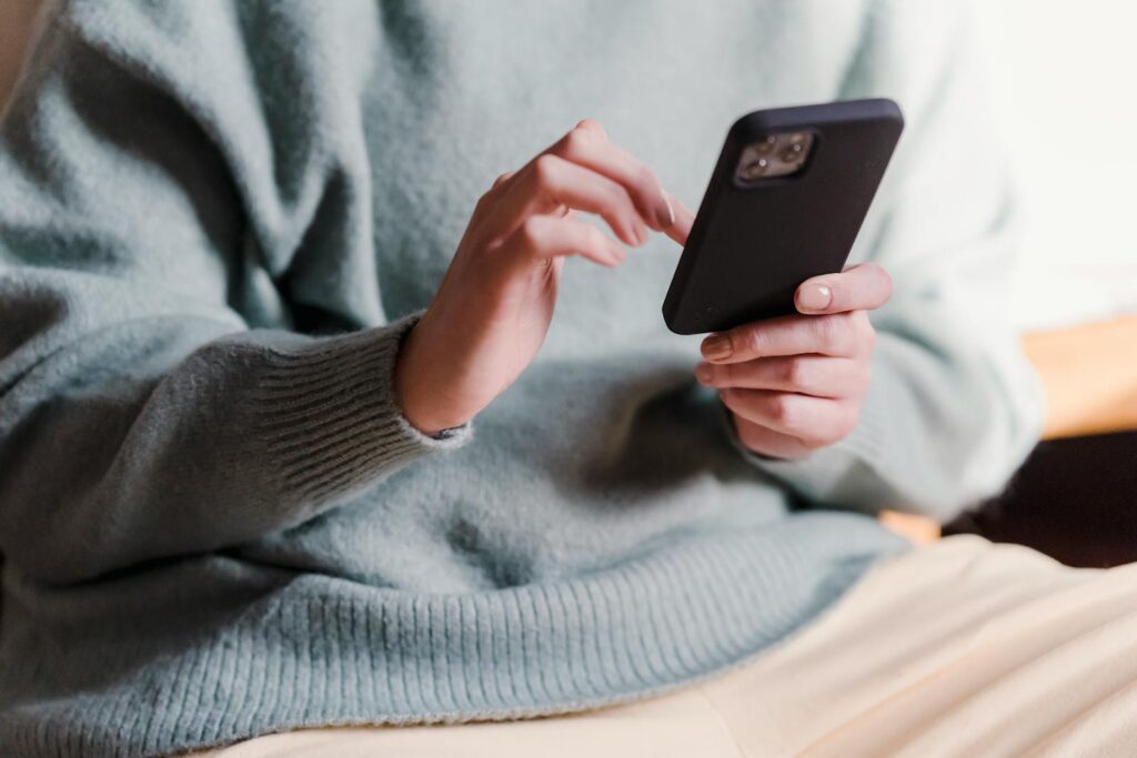 Crop faceless female in casual clothes using smartphone and touching screen while chilling at home