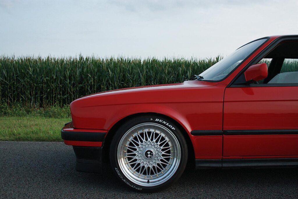 Side view of a classic red car with alloy wheels on a rural road beside a cornfield.