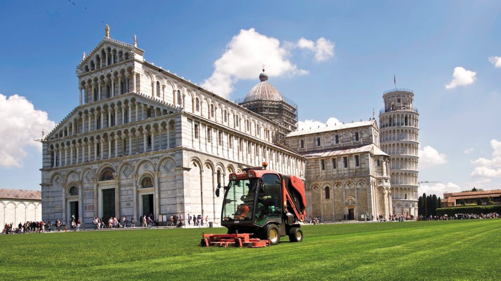 A lawnmower working near Pisa Cathedral and Leaning Tower on a sunny day.