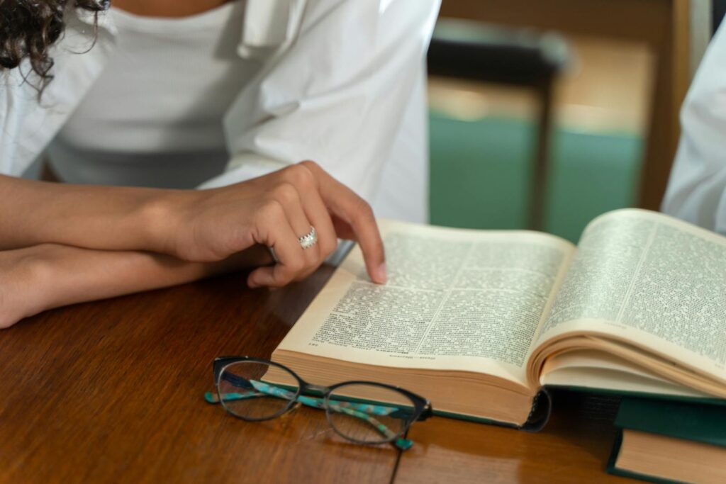 Close-up of a person reading an open book with eyeglasses on a wooden desk.