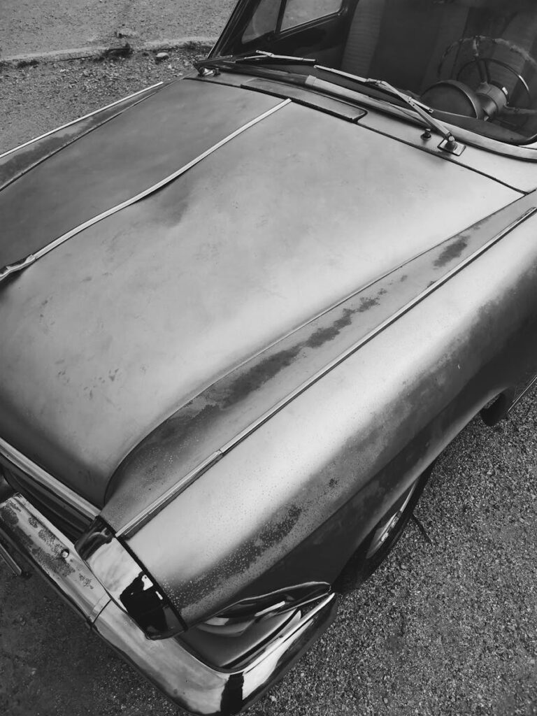 Close-up of a vintage car with a rusty, dented hood in black and white.