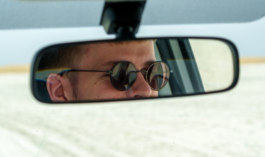Man wearing sunglasses reflected in car's rearview mirror.